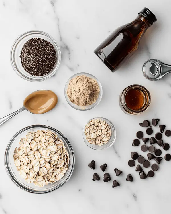 Ingredients for no bake lactation cookies including oats, flaxseed, brewer's yeast, peanut butter, honey, vanilla, and chocolate chips arranged on marble counter